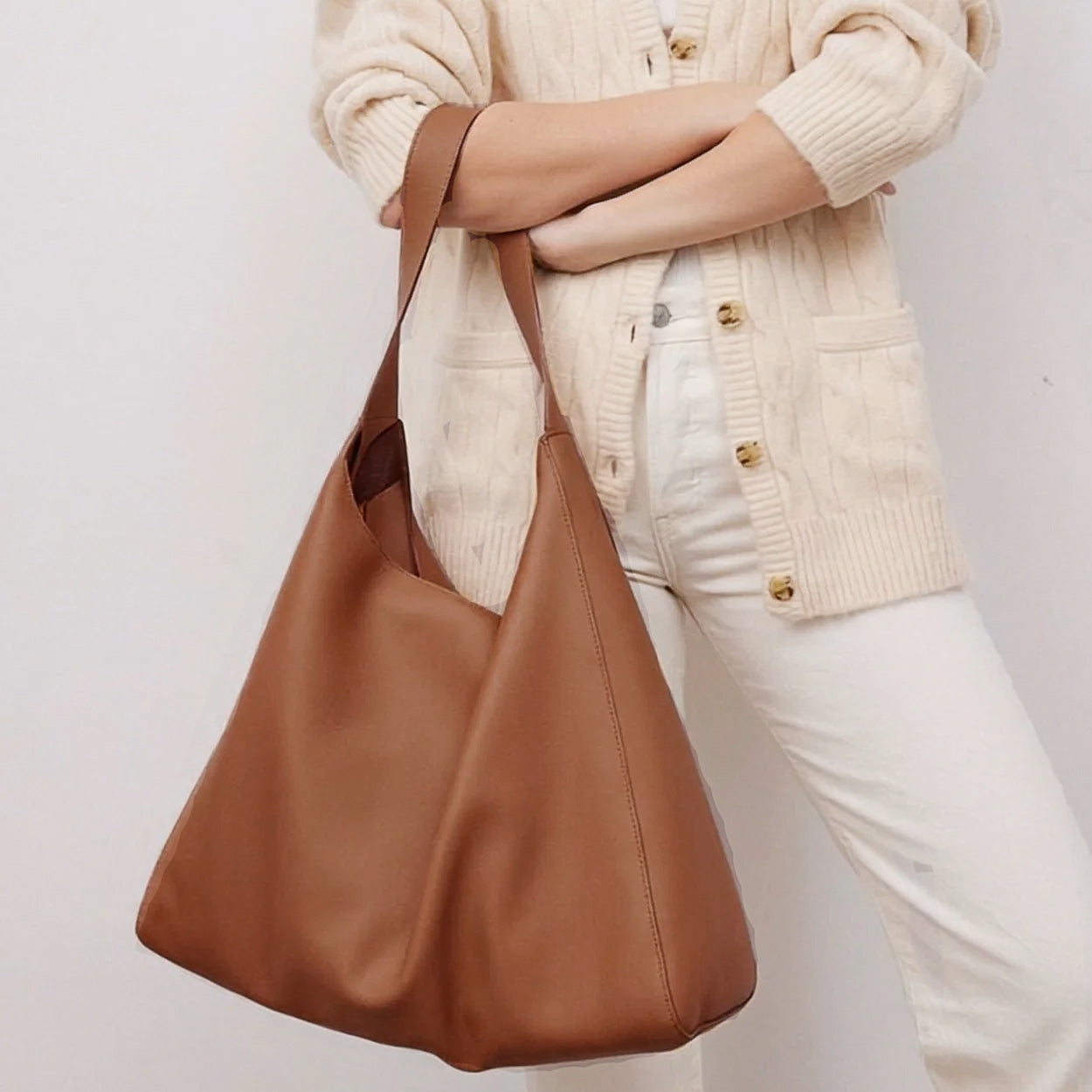 Brown leather handbag held by a person wearing a beige sweater and white pants on a light background - Saddle