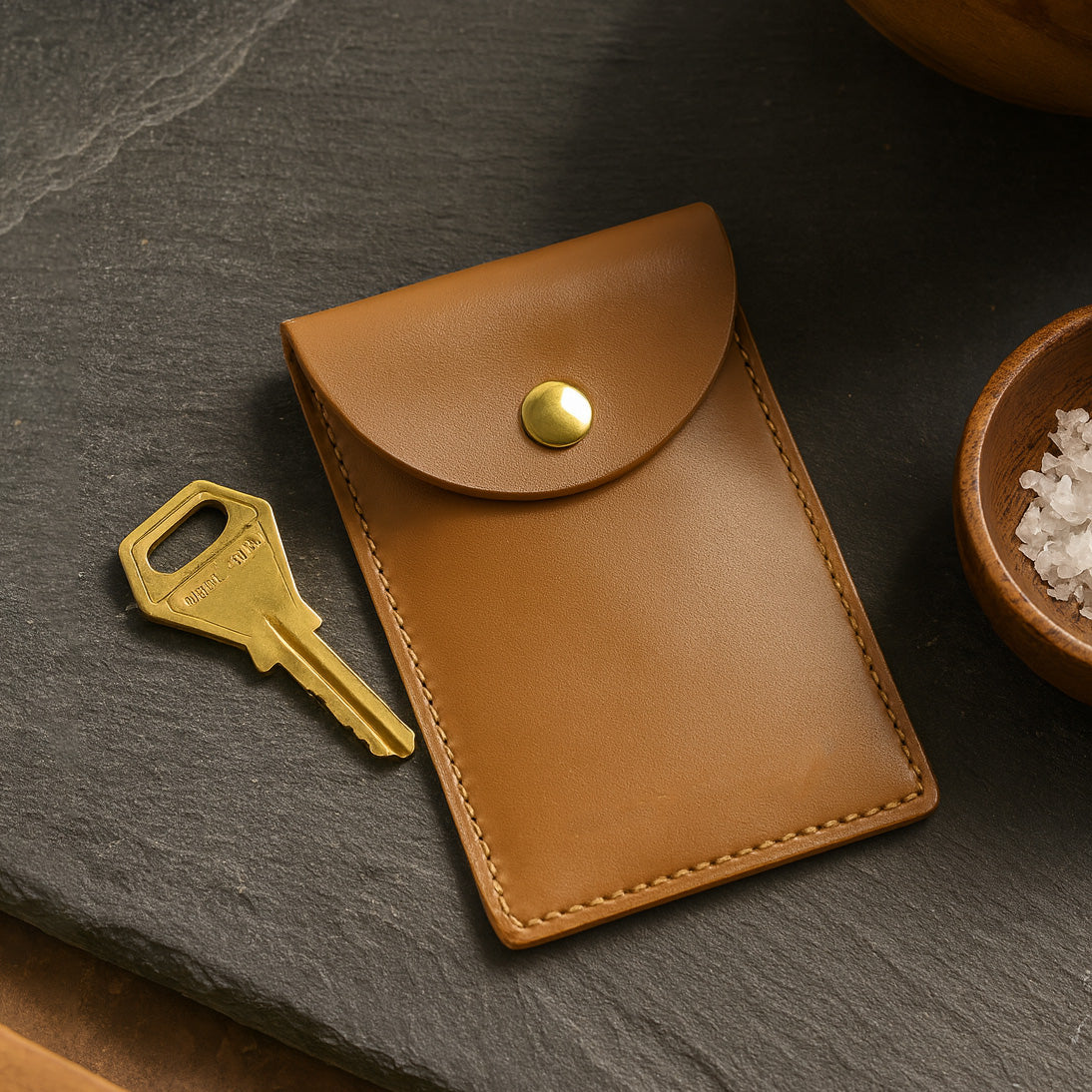 Brown leather pouch with a gold button and key on a dark surface - Saddle