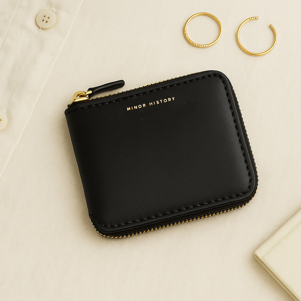 Black wallet with gold zipper and 'Minor History' branding on a beige surface. - Black