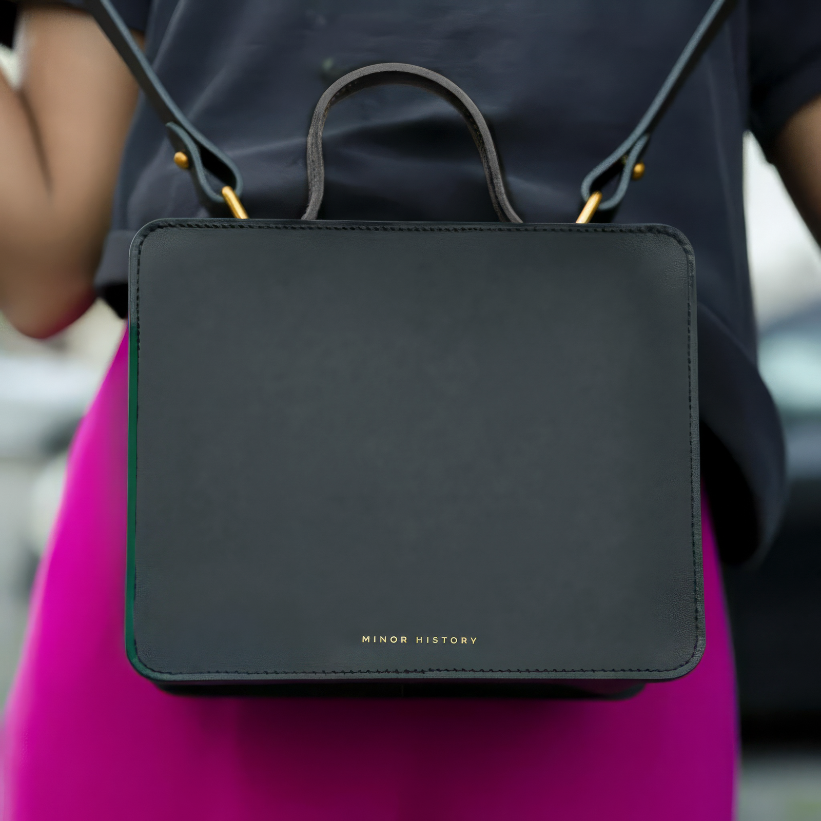 Black handbag with 'Minor History' branding held by a person wearing a dark shirt and pink pants. - Black