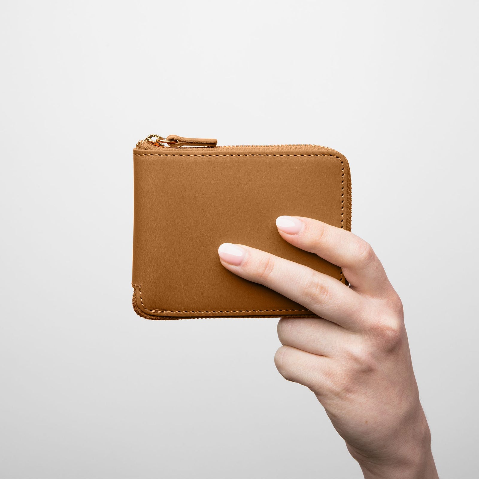 A hand holding a brown leather wallet with a zipper closure. - Saddle